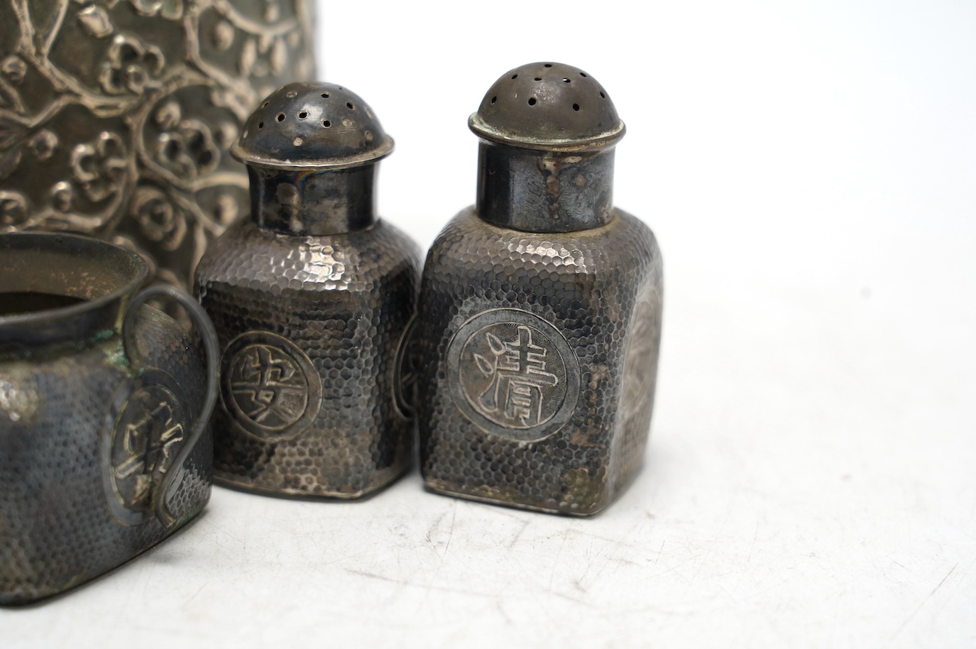 A Chinese embossed white metal cylindrical jar and cover, 11.2cm, and a similar white metal four piece cruet set. Condition - poor to fair